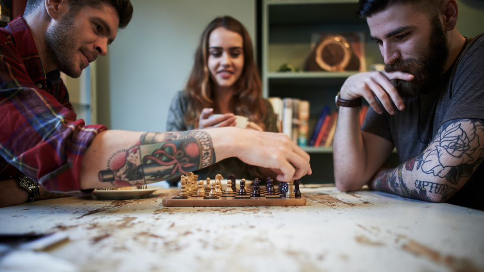 Two tattooed young men playing chess while a female watches, drinking a mug of tea.