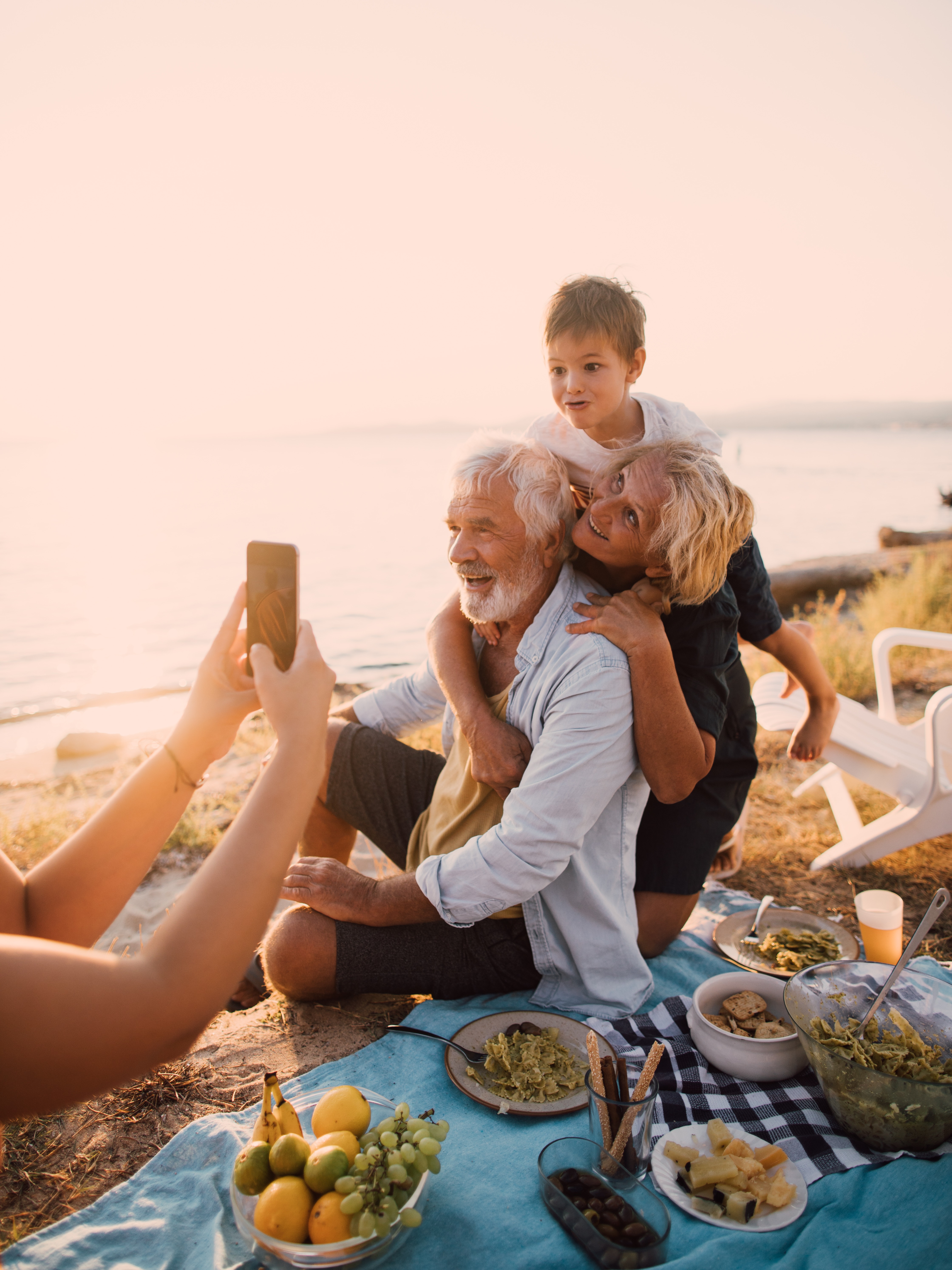 Elderly couple and young boy smiling for a photo during a seaside picnic at sunset.