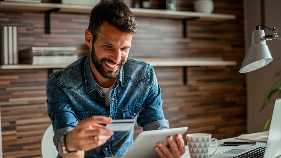 A man using a tablet at a home office, holding a credit card.