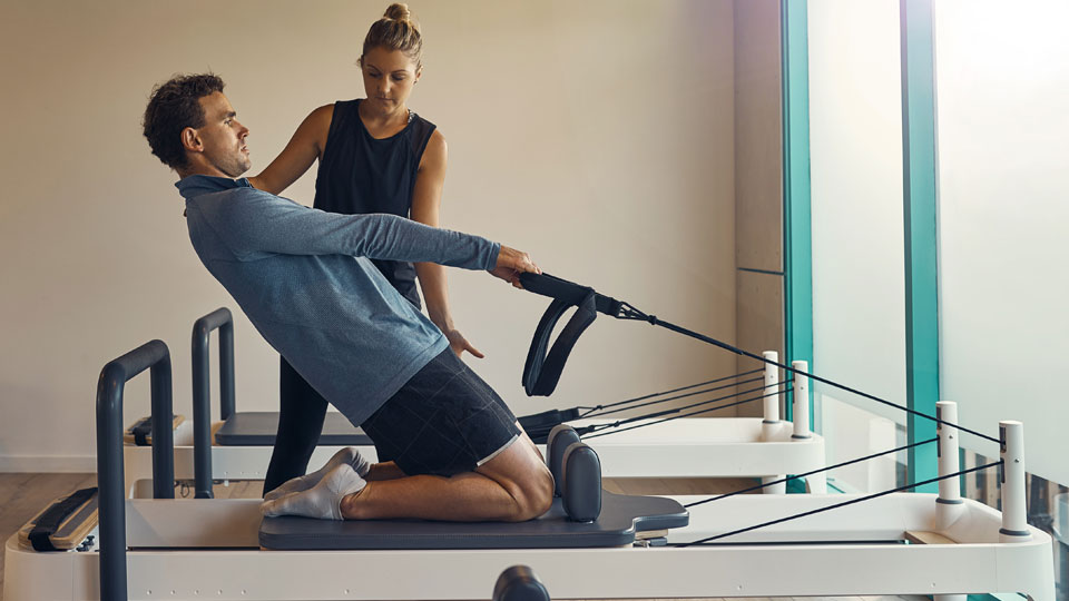 A woman instructs a man on using a pilates device