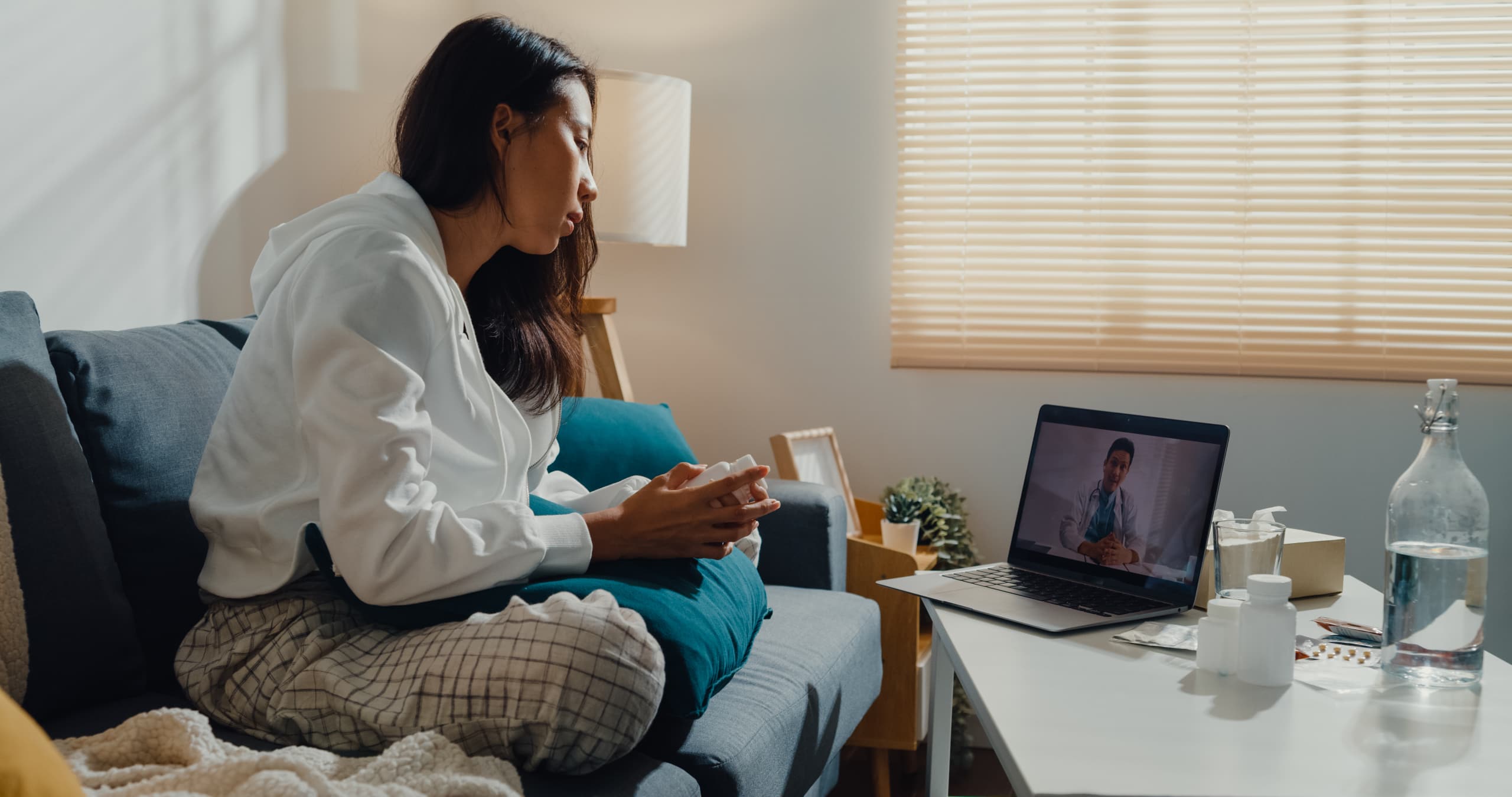 A sick young person using a laptop talk to doctor for a telehealth appointment from the living room in their home.