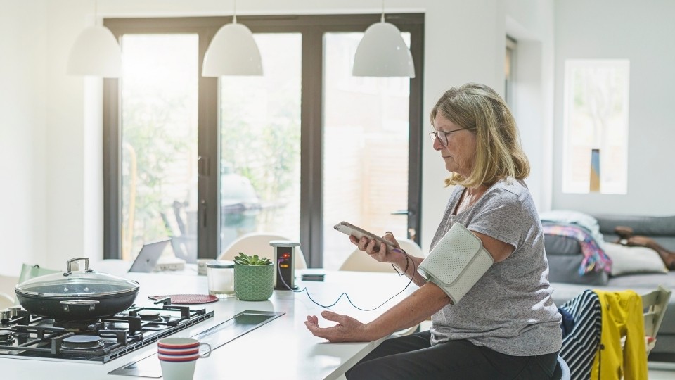 A woman taking her blood pressure at her kitchen bench.