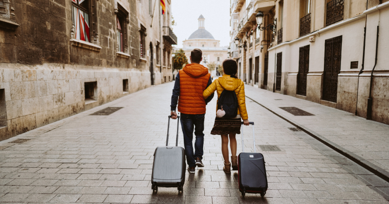 A man and a woman walking through a European street with their suitcases being wheeled behind them