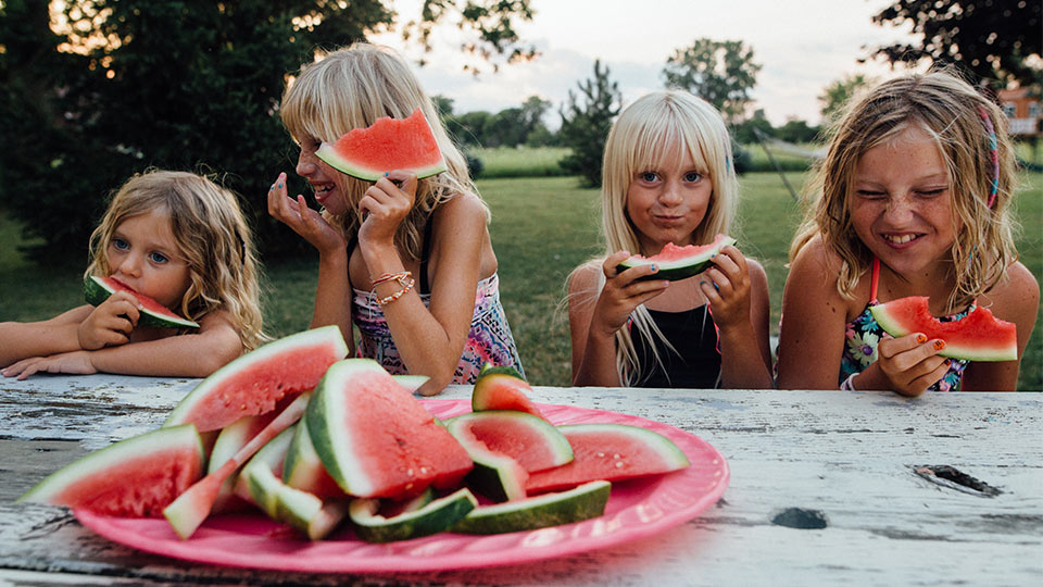 Four children standing at a table outdoors eating watermelon.