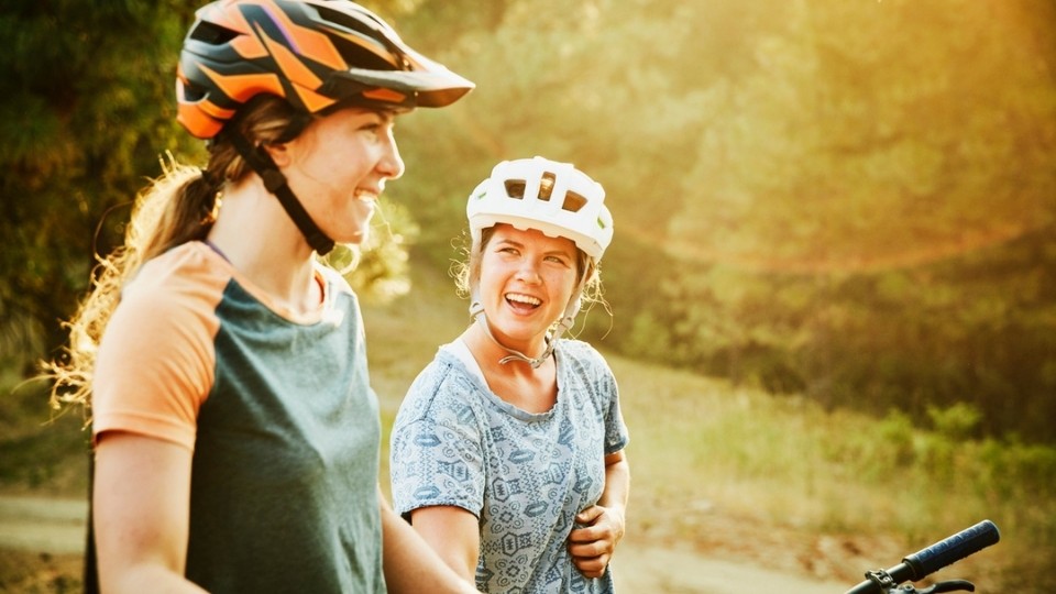 Two women wearing bicycle helmets laughing as they stop riding for a break.