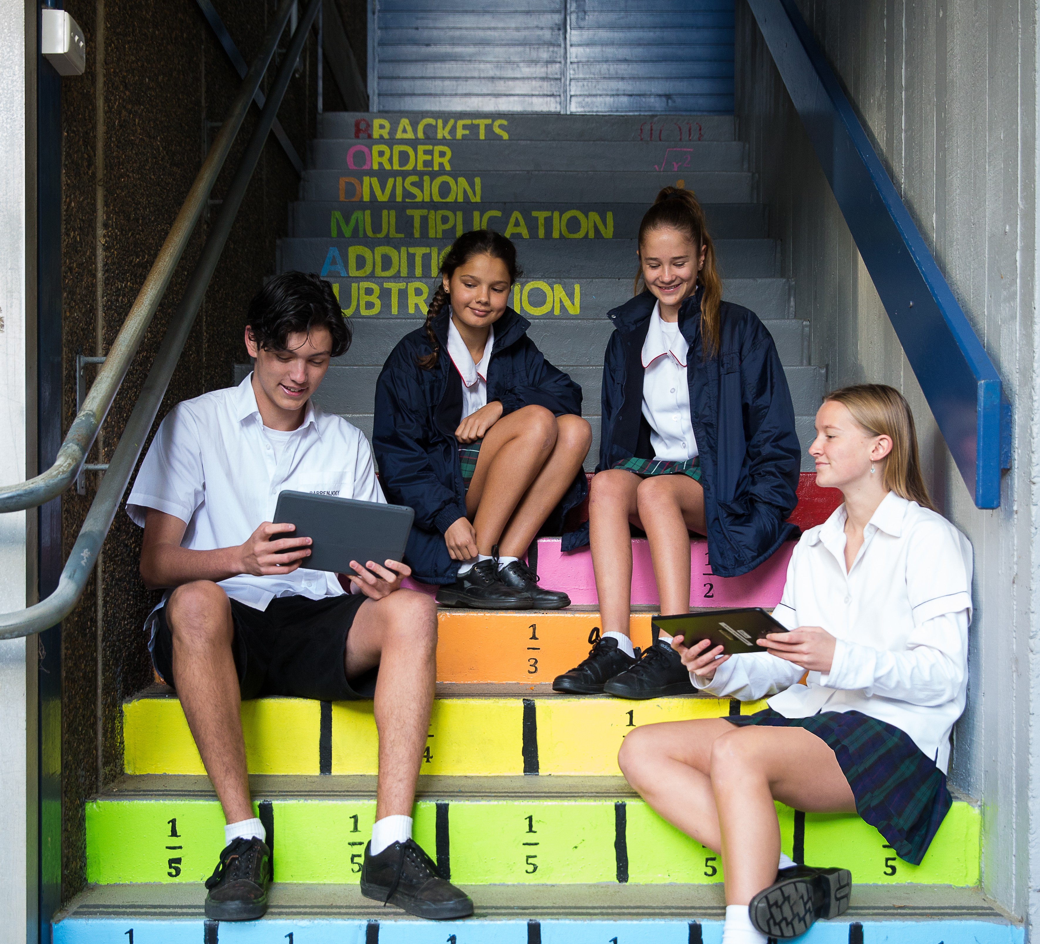 Four students sitting on colourful steps in a school setting looking at their tablets and talking