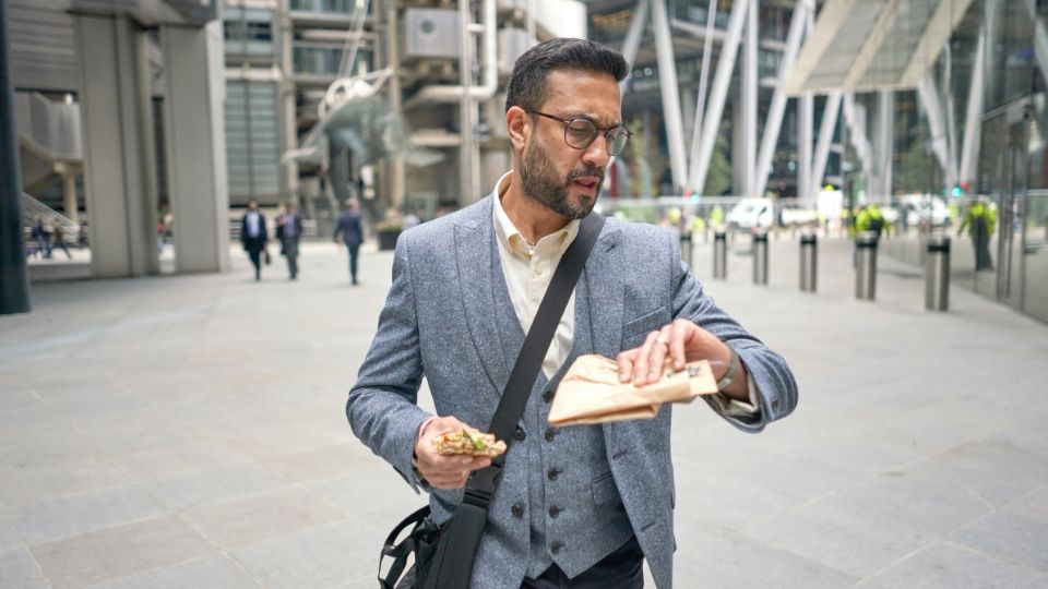 Man walking from the office holding a sandwich and checking the time on his watch.