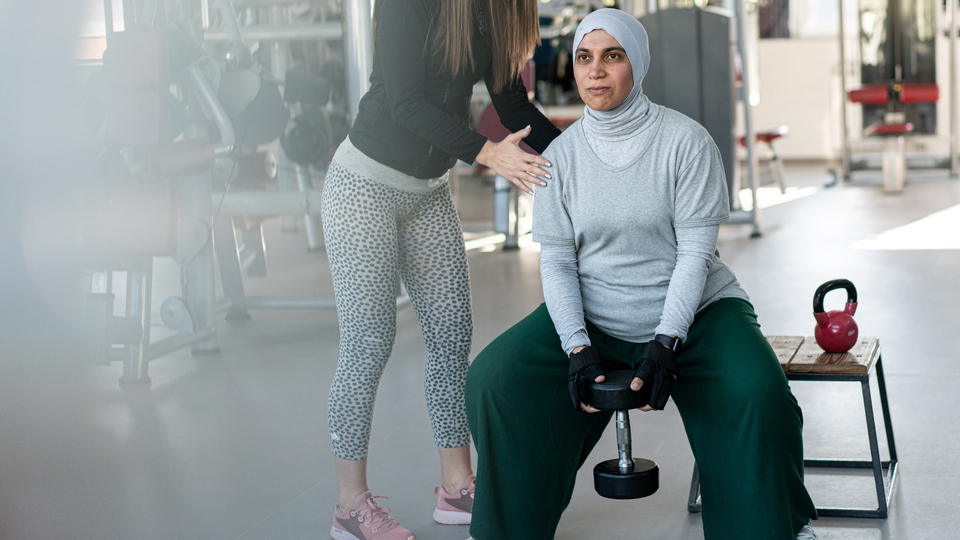 A women lifting hand weights at the gym with the support of another person.