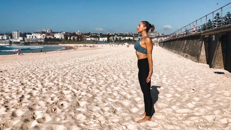 Cassey Maynard taking a big breath in with her arms down by her side as she stretches on the beach.