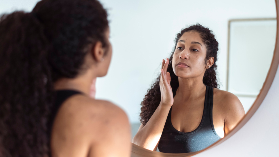 Middle-aged woman with dark, curly hair looking into a mirror at her skin.