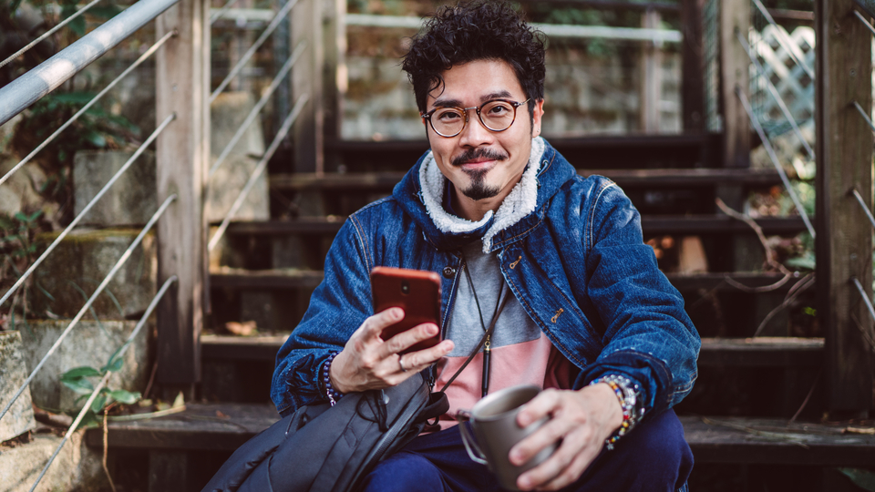 Young person with beard and glasses holding his phone and a mug, sitting on stairs.