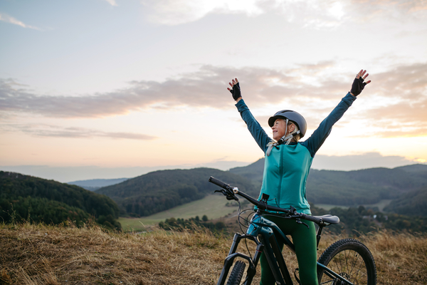 A woman on a bike wearing a helmet at sunset, with her arms up in the air