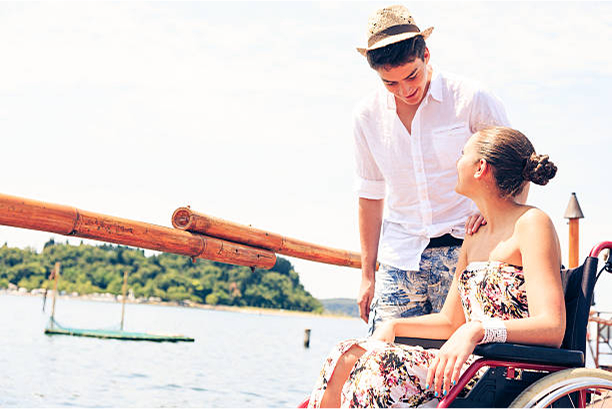 A woman in a floral dress sits in a wheelchair, smiling up at a man in a white shirt and straw hat. They are in front of a scenic body of water. 