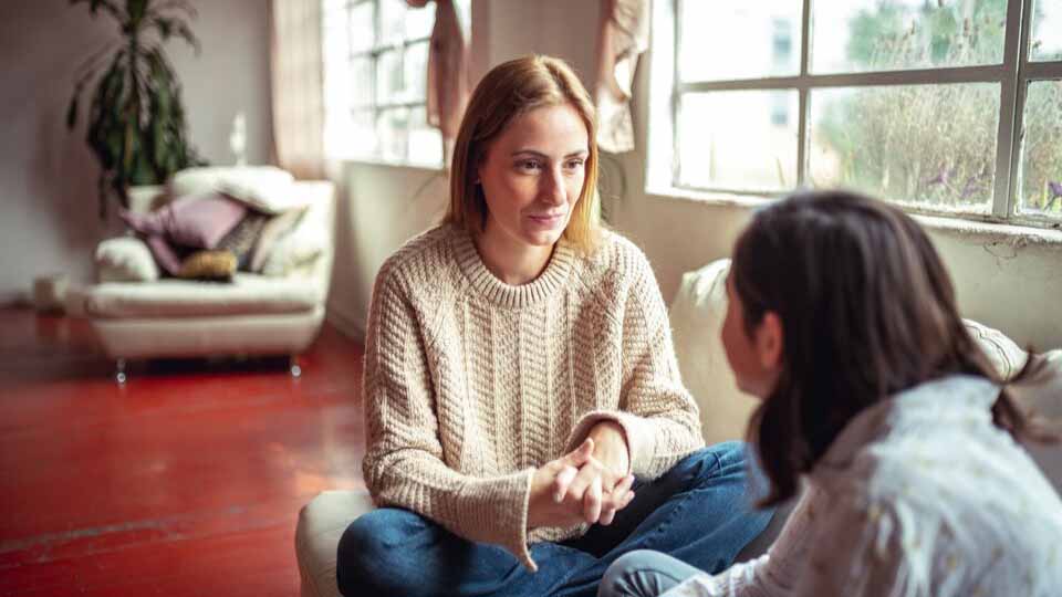 Two women sitting cross-legged on a lounge, talking to each other.