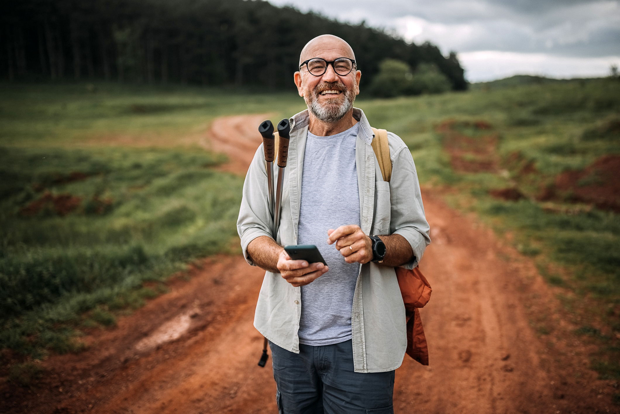 A senior man hiking in Serbia, along a red dirt road, holds his phone while smiling at the photographer