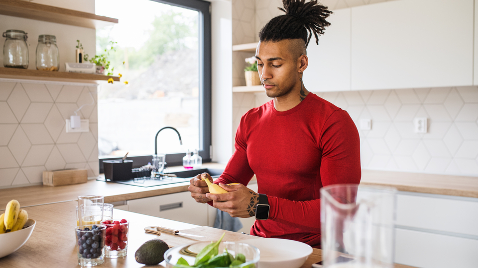 Man standing in kitchen peeling a banana with other fruits and vegetables prepped.