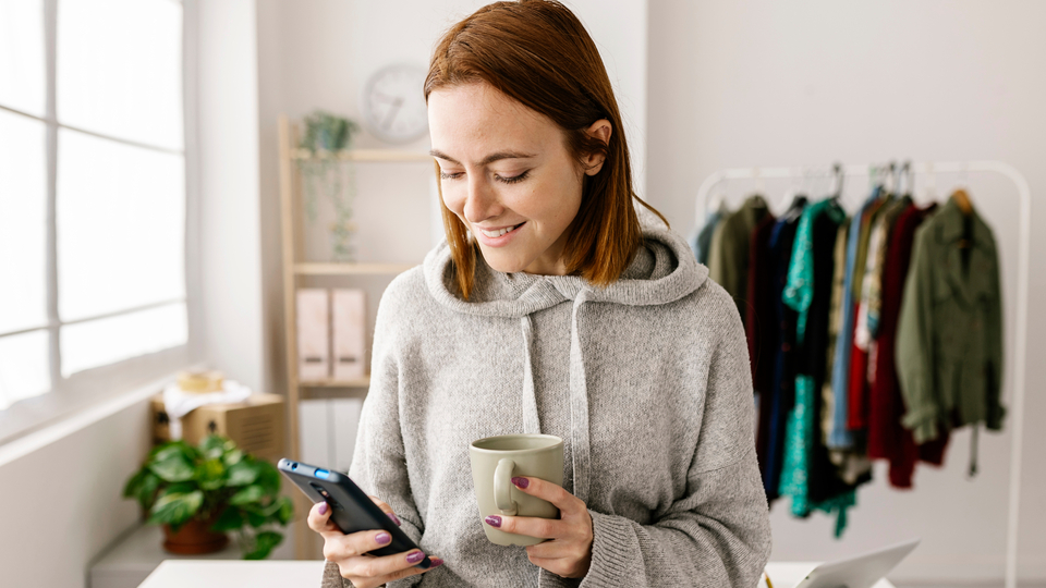 Young woman with red hair standing in a minimalist room, holding a mug and looking at her smartphone.