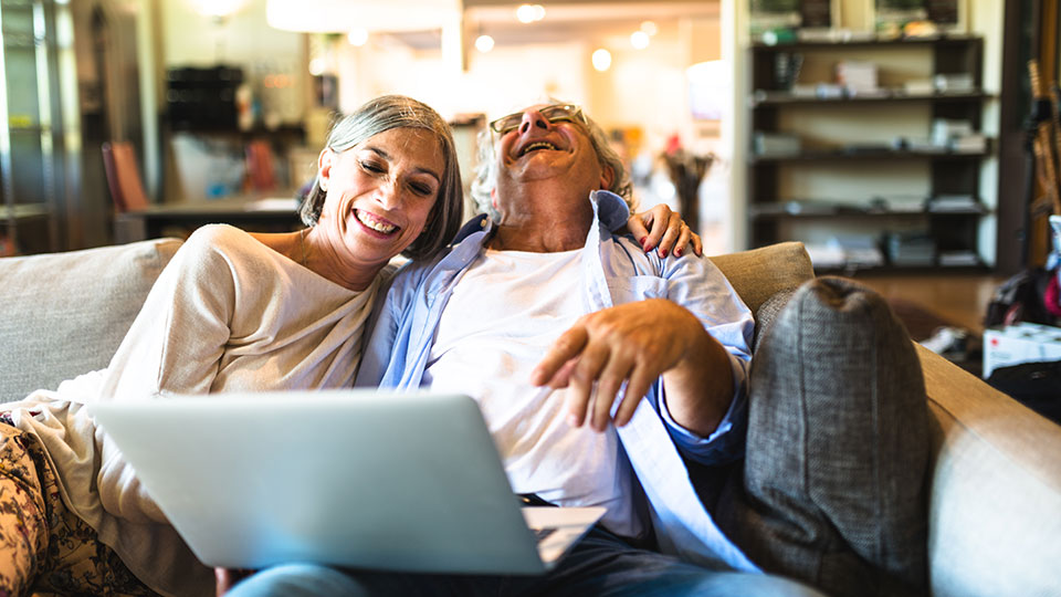 Senior couple sitting on their lounge, looking at a laptop while smiling.