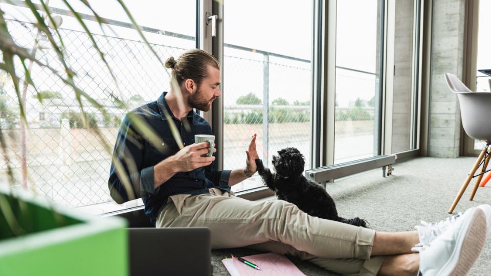 A young man holding a coffee sits on the floor and high fives his dog