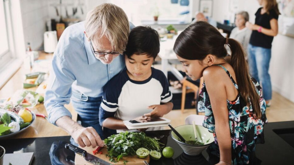 Two children sitting on a kitchen benchtop, a man stands near them chopping up vegetables while they look on.