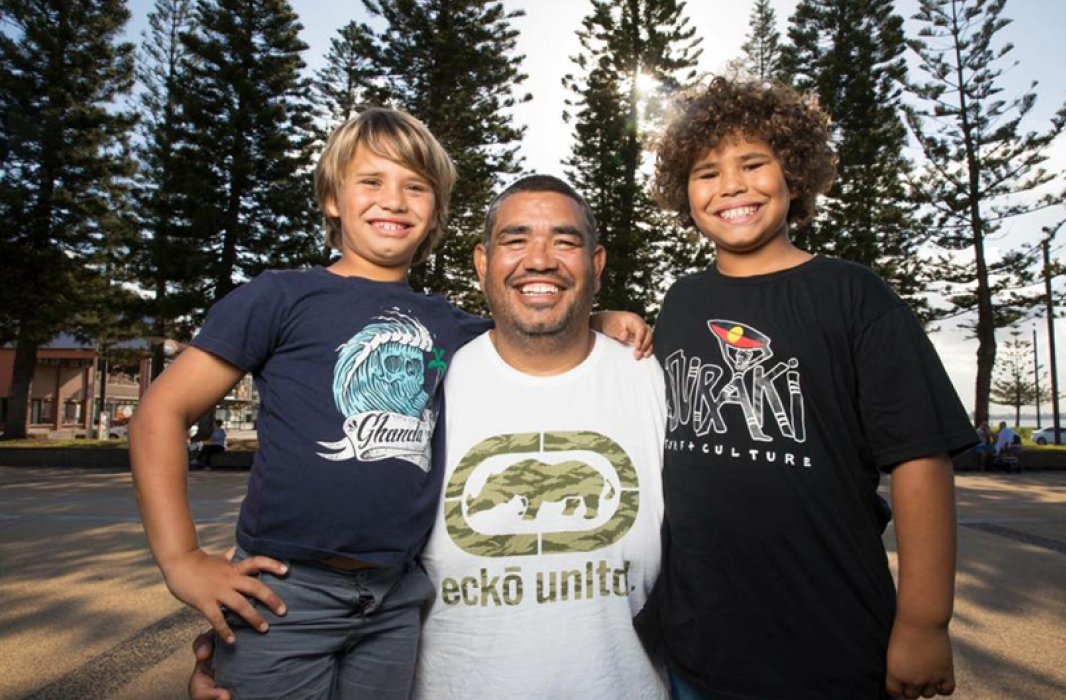 A smiling man with two boys standing outdoors with tall pine trees and sunlight behind them in a park setting.