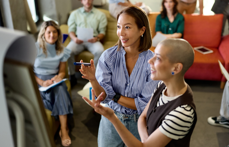 Happy creative women reading plans on whiteboard while leading a business presentation with their colleagues at casual office. Focus is on Korean woman.
