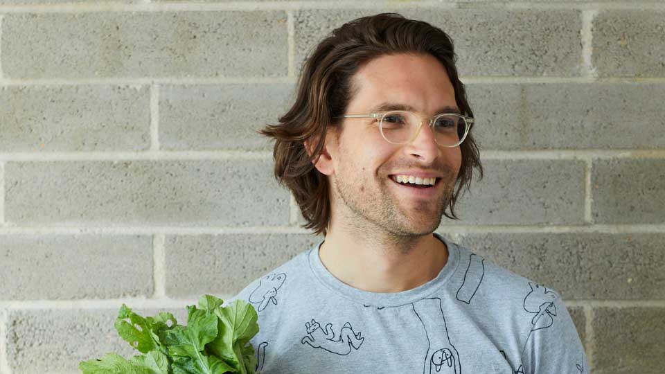 Close up photo of a man standing against a grey brick wall holding a bunch of vegetables, smiling as he looks into the distance.