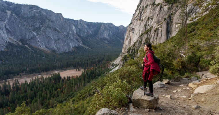 A woman in hiking boots and clothes standing on a rock overlooking a river in front of a mountain