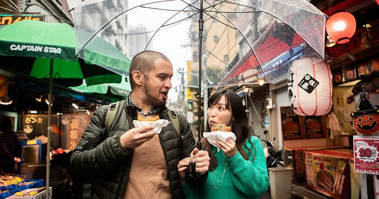 A man and a woman eating food from markets, walking under an umbrella