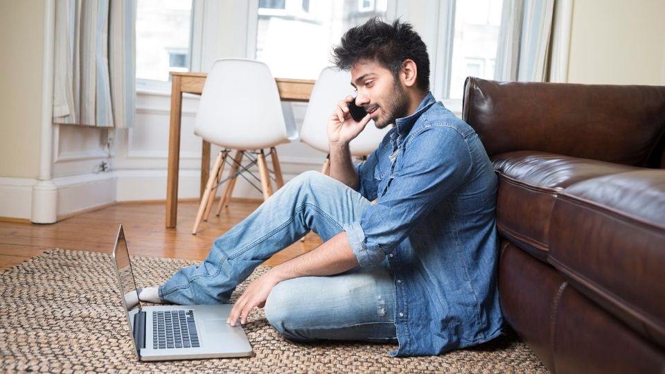 Man sitting on the floor of his living room using a laptop while on the phone.