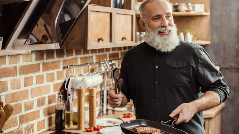 A middle-aged bearded man cooks up meat in a pan in the kitchen.