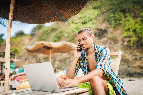 A digital nomad sitting under an umbrella on the beach on a phone call and working in front of a laptop