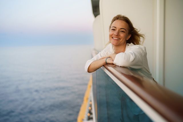 A woman on a cruise ship leaning on a railing and looking out at the ocean at dusk