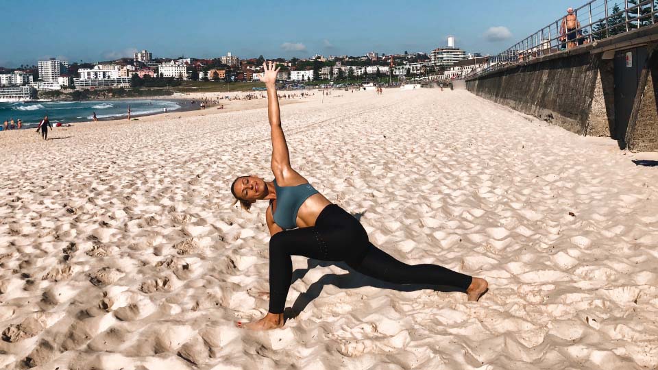 Cassey Maynard doing a yoga stretch at the beach with her knee lunging forward and her body twisted open to the side.