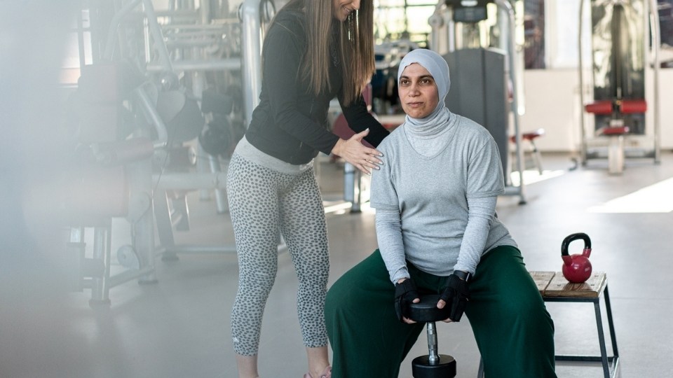 A woman wearing a Hijab holding a weight while being instructed by a female personal trainer.