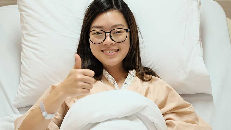 A young woman in a hospital bed gives the thumbs up.