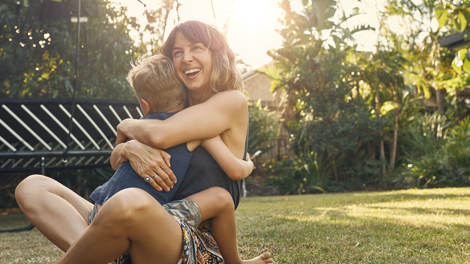 Mum in her early 30s sitting in the backyard and hugging young son.