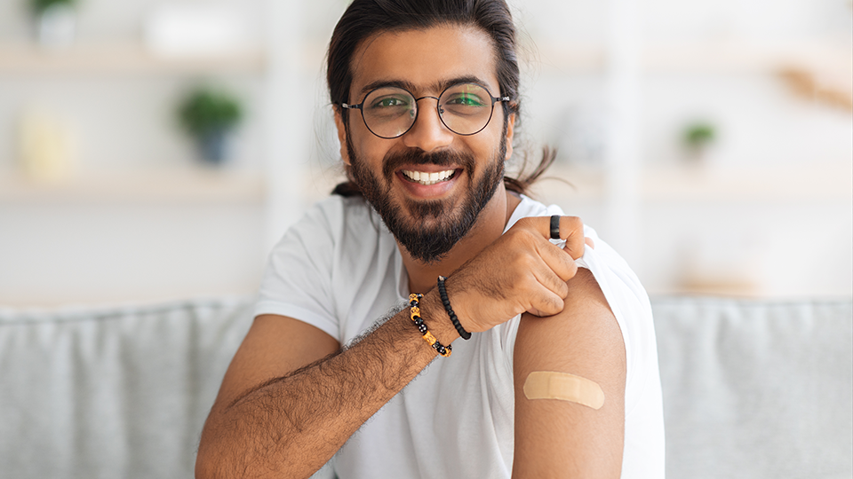 Smiling man lifting his t-shirt sleeve to reveal a bandaid on his arm at the site of a vaccine injection.