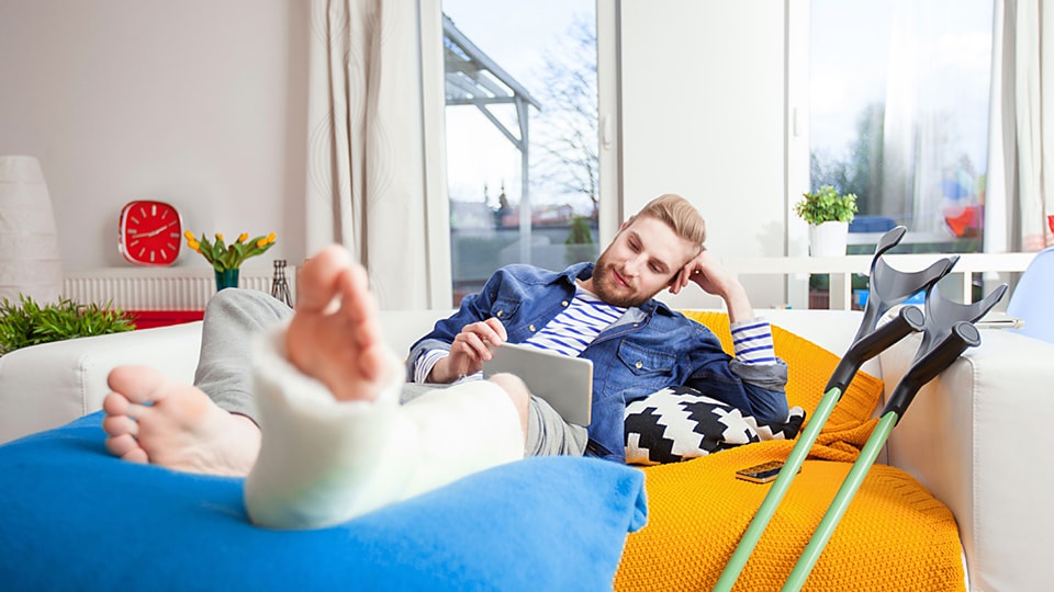 Man lying on a lounge, looking at an iPad with his leg outstretched in a cast and crutches next to him.