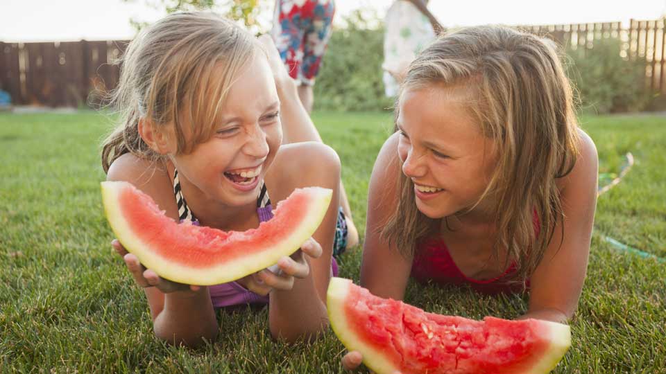 Two girls laying on grass eating big pieces of watermelon.