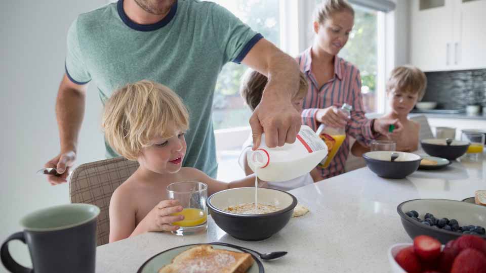 A family at a breakfast table with a father pouring milk on his son's cereal.