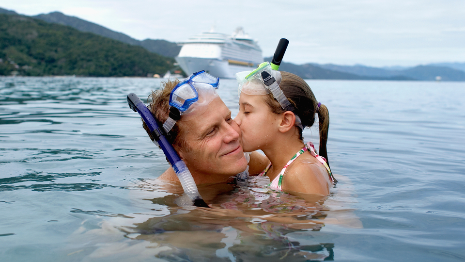 Dad and daughter wearing snorkels in the ocean. The daughter is giving her had a kiss on the cheek. Cruise ship in the background.