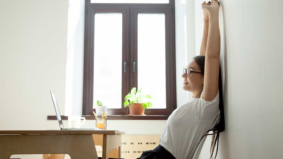 A woman smiles as she stretches back from her laptop screen with her arms in the air.