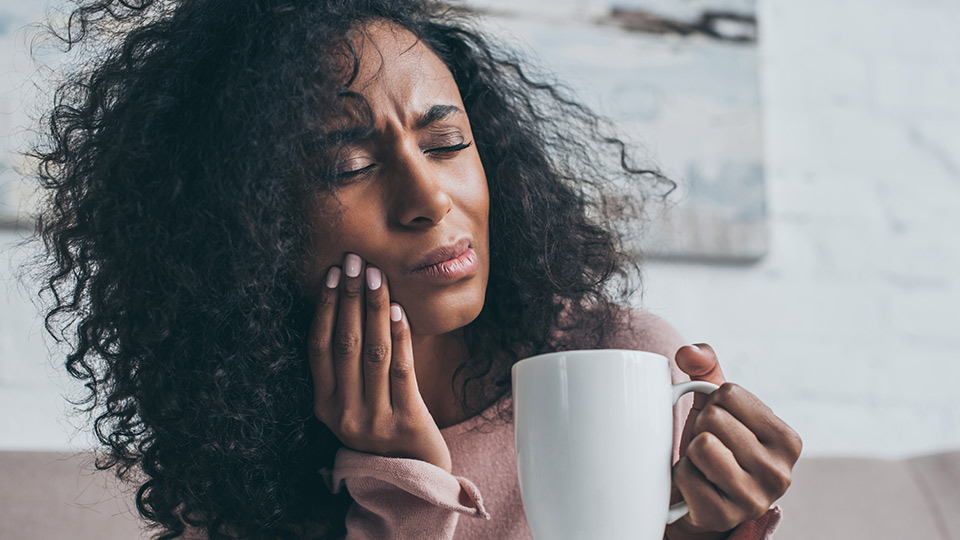 A woman holding her jaw due to a toothache with one hand and a cup of coffee in the other.