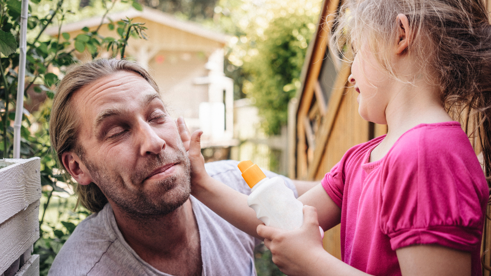 Hipster dad getting sunscreen applied by his 5-year old daughter who's wearing a pink shirt.