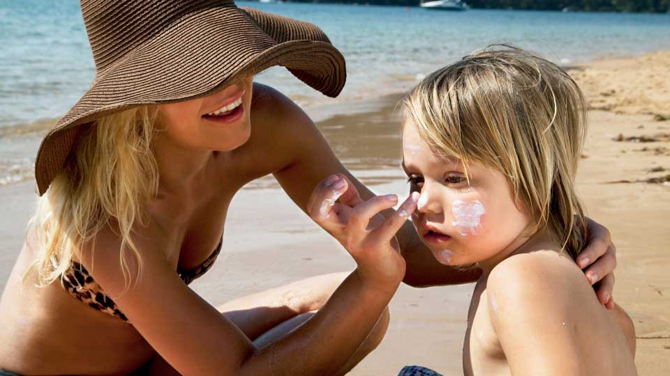 Woman applying sunscreen to a toddler's face at the beach.