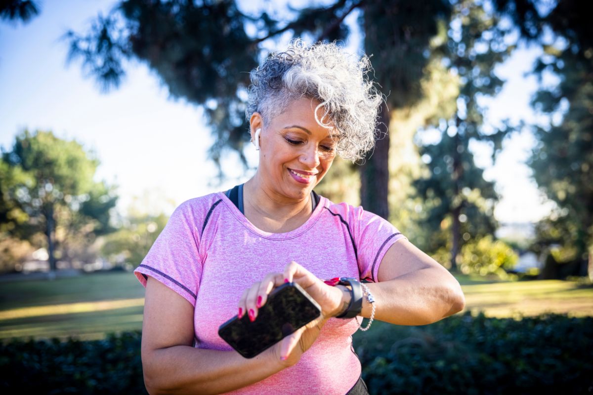 Senior woman running in a park while wearing a fitness tracker, surrounded by trees and greenery.