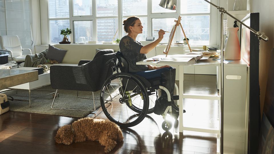 A woman in a wheelchair painting with her dog lying nearby.