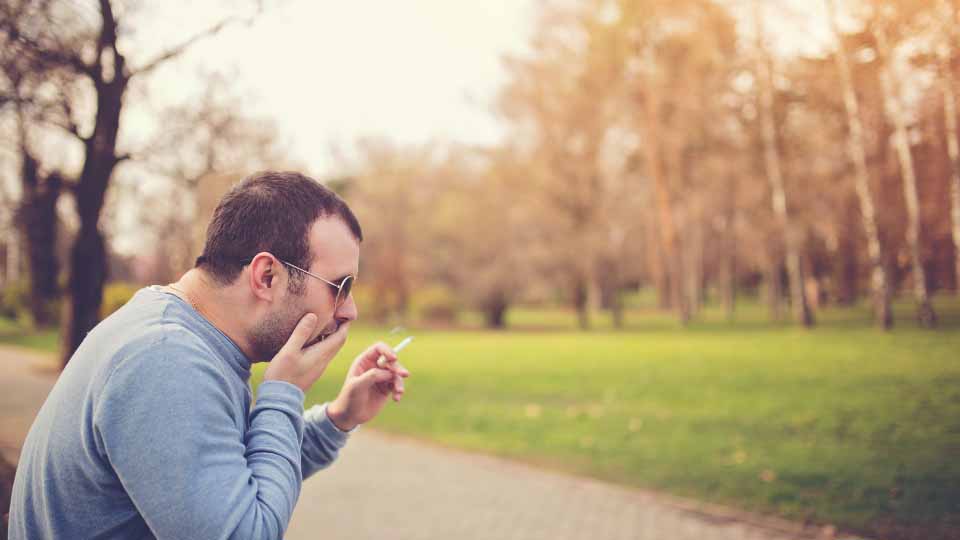 A man smoking in a park covers his mouth to cough.