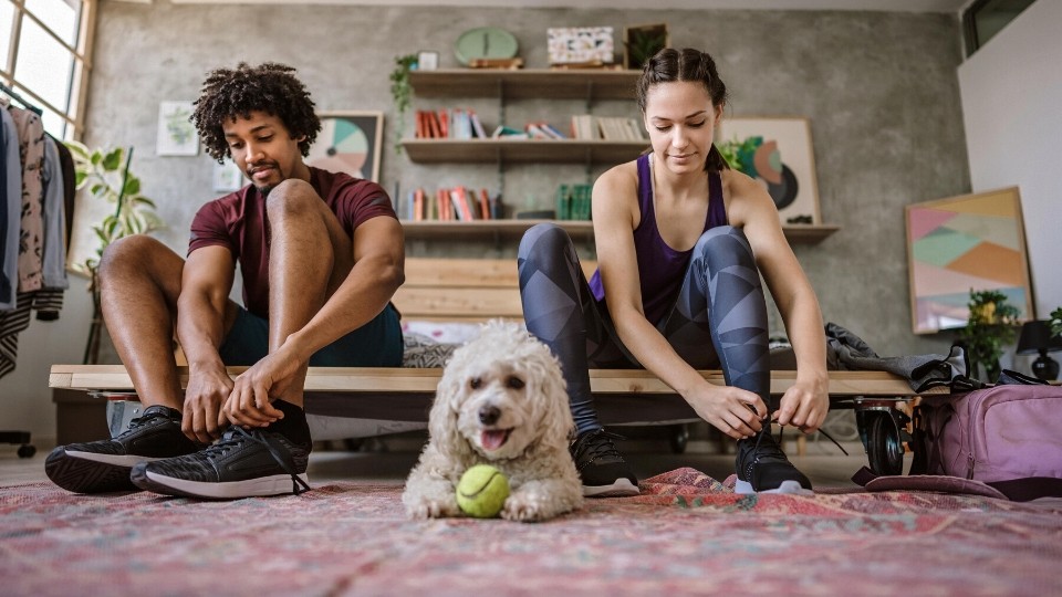 Two people tying the shoelaces on their running shoes, a dog with a tennis ball sits in between them.